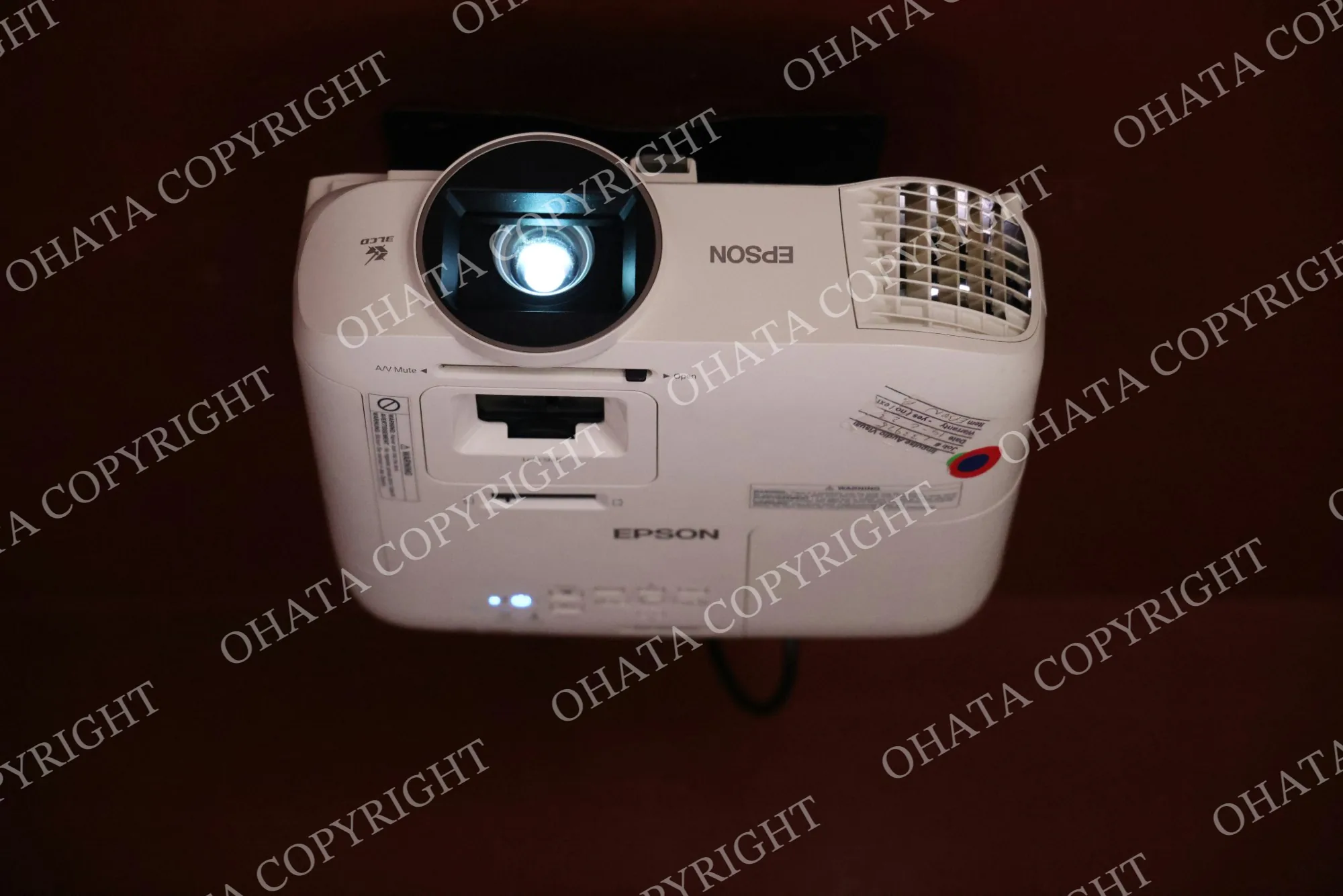 White Epson video projector installed upside down with its projection lens illuminated.