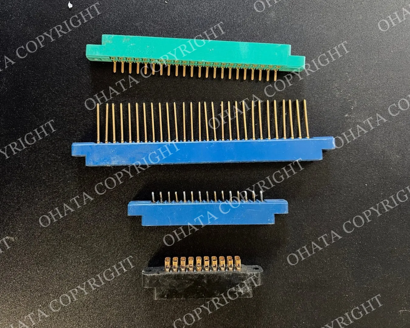 Four loose plastic connectors in green, blue, and black housings arranged on a dark surface, showing different sizes, pin counts, and gold-colored contact designs.