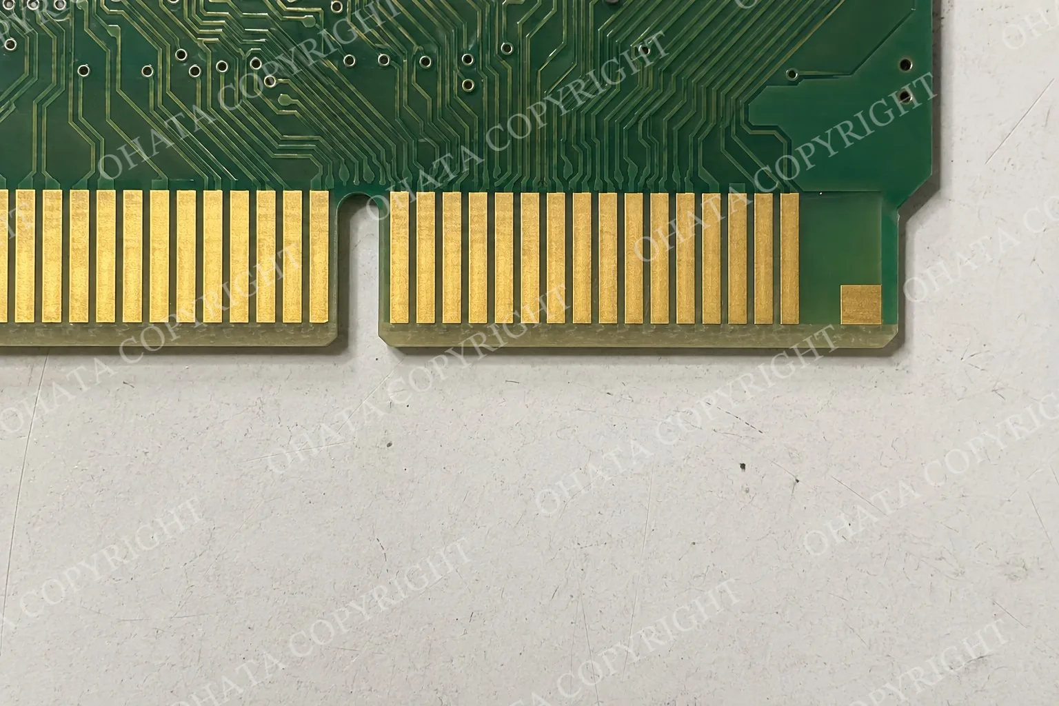 Top-down close-up of a circuit board edge with gold fingers, an alignment notch, a beveled edge, and an intentionally shorter contact on a white-gray worktable.