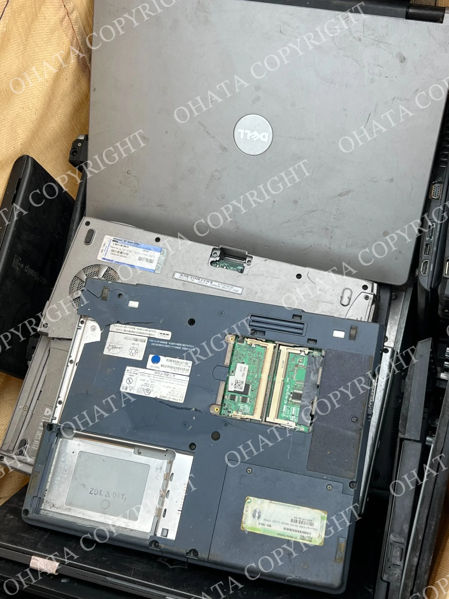 Stack of used Dell laptops, including one turned over with bottom covers removed and memory modules exposed.