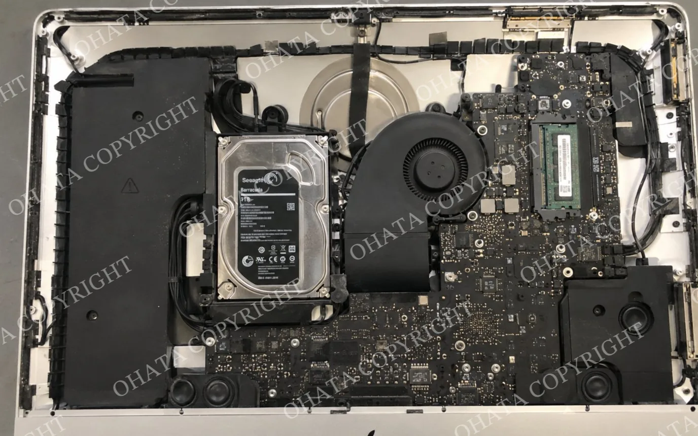 Interior of an opened Apple iMac showing the logic board, hard drive, cooling fan, memory module, speakers, wiring, and internal chassis.